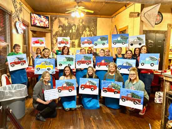 A photo of artist Deb Brown at her first ever painting party that launched her business, Art & Grace. She stands with a group of smiling women holding up freshly done canvas paintings.