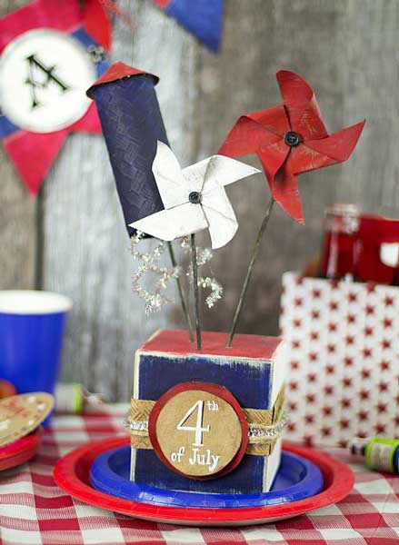 A 4th of July centerpiece made of a wooden block painted read and blue with some pinwheels sticking out of the top and tied with ribbons