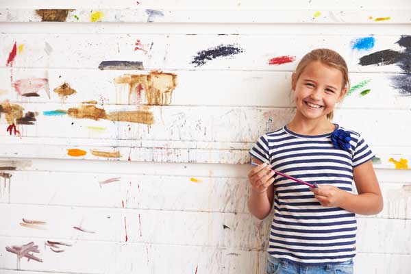 A young girl smiling and holding a paintbrush in front of a blank white wall.