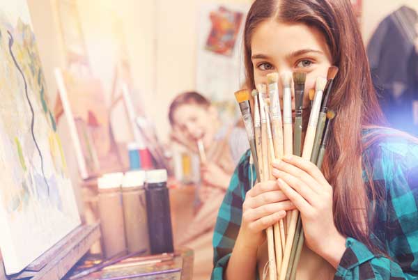 A girl painting on canvas looks towards the camera and holds up and assortment of paintbrushes with a smile.