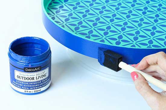 Someone paints an outdoor end table using a vivid blue outdoor living acrylic paint from DecoArt