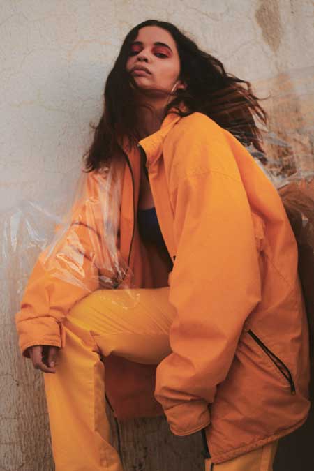 A girl in a marigold yellow tracksuit leans against a wall.