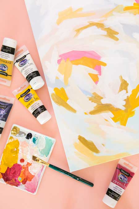 An abstract painting is painted using marigold yellow, white, and pink for a free-form and fun look. Bottles of acrylic paint are scattered around the painting.