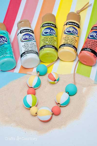 A line of Americana Acrylic paint bottles in coastal, beachy colors lined up to a painted beach ball necklace on a pile of sand.