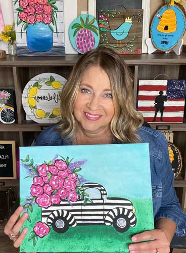 Artist Deb Brown holds up an acrylic painting of a truck with flowers in the truck bed, she's smiling.