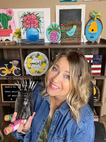 Artist Deb Brown smiles while she holds a bottle of DecoArt acrylic paint and some paintbrushes.