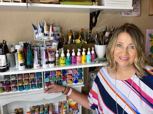 Artist Deb Brown stands in front of shelves of DecoArt acrylic paint with a smile on her face.