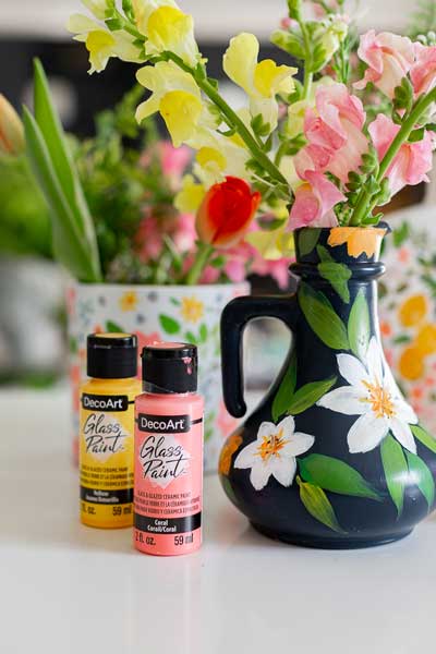 DecoArt Glass Paint in yellow and pink is sat next to a glass vase painted using the acrylic paint in a fun floral design. The vase is filled with flowers for Mother's Day.