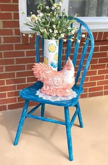 Farmhouse style blue chair with a painted chicken figure sit on a front porch.