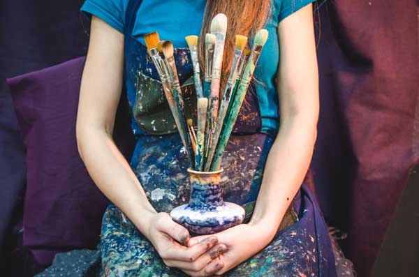 A woman in a paint-splattered apron holds a vase filled with a variety of messy paintbrushes