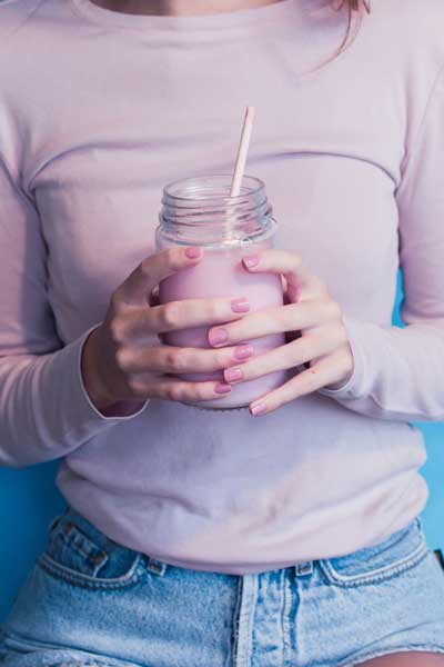 A woman wearing a light pink sweater holds a mason jar with a pink fruit smoothie in it