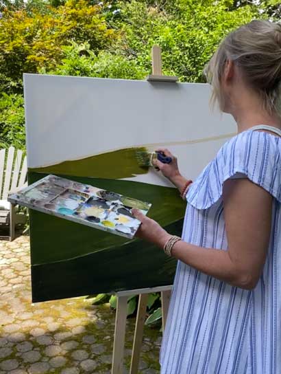 A woman paints a landscape with her back to the camera.