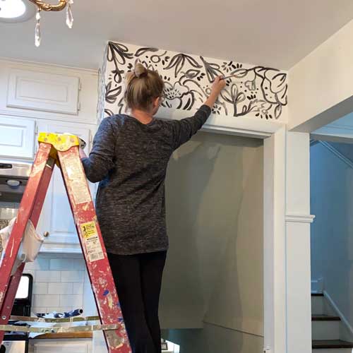 Designer Jennifer Rizzo paints a black and white line art mural above the doorway leading into a kitchen.