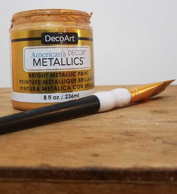 A bottle of DecoArt's DIY paint Americana Decor Metallics in gold sits on top of furniture about to be painted.