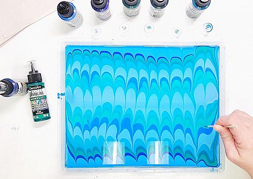 A water marbling artist uses a stick to create an arches pattern in the water marbling bath. The pattern is in ocean colors.