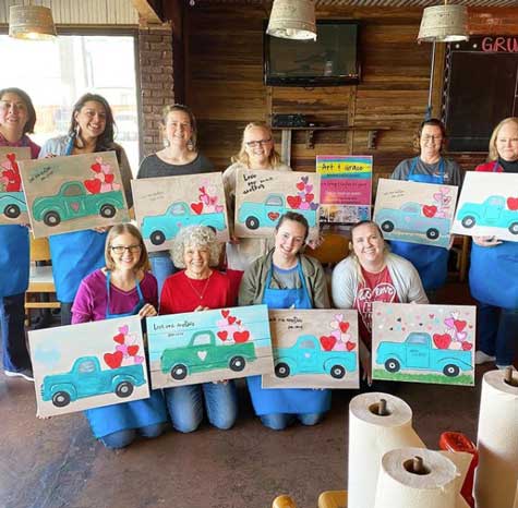 A group of smiling women hold up canvas acrylic paintings they just completed during a paint class.