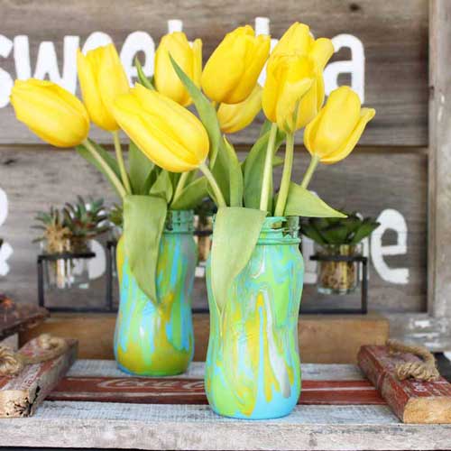 Mason jars are decorated with an acrylic paint pour and filled with yellow tulips as a springtime centerpiece 