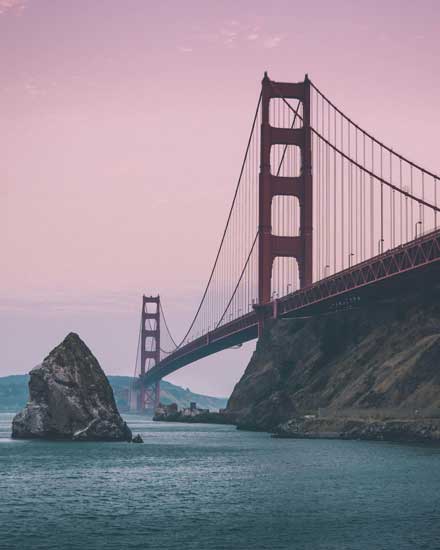 The sun sets over the holden gate bridge, the pink sky contrasts against the cool blue water and red bridge.