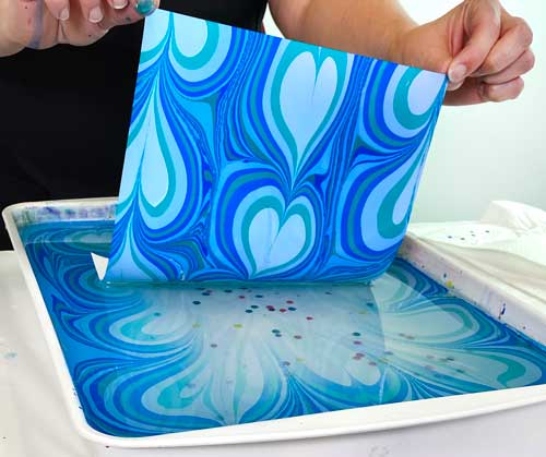 A person lifts a paper off of a water marbling bath to revel a blue heart design.