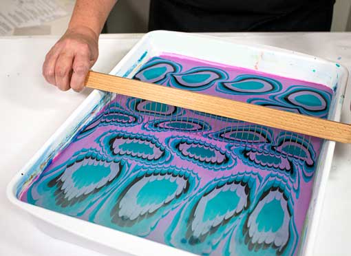 A rake is being pulled through a water marbling bath to create an intricate pattern.