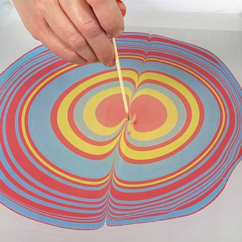 A person drags a stylus through a water marbling pool to create a swirled design.